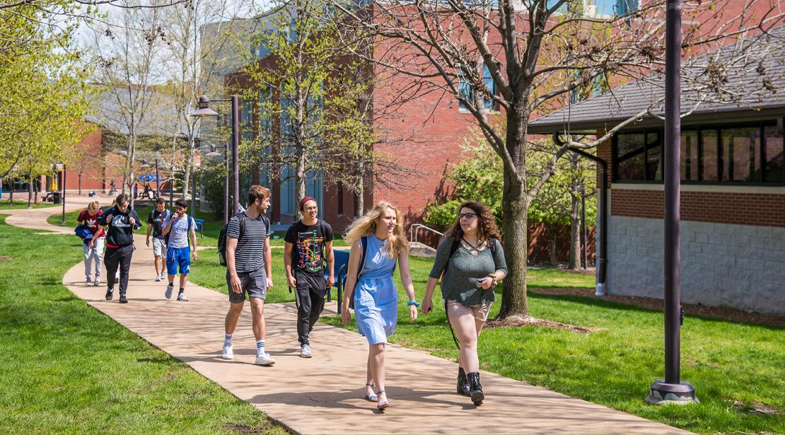 Students walking a brick-layered sidewalk under trees beginning to leaf in early spring.