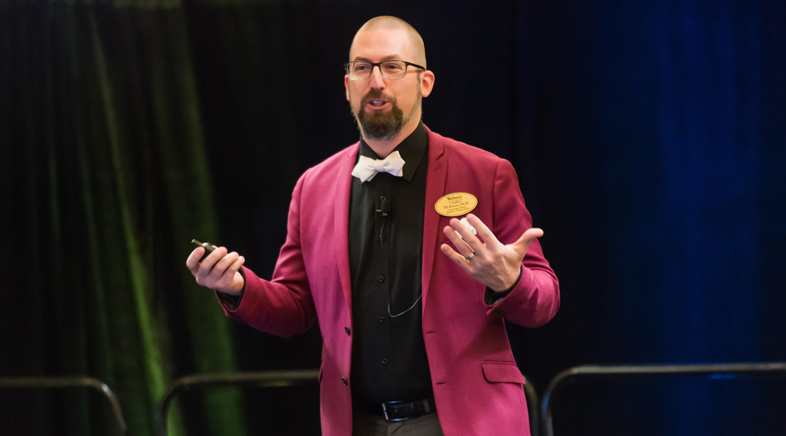 DJ Kaiser, a bald, light-skinned man with a dark brown goatee and glasses wearing a magenta jacket over a black shirt with a white bowtie, gestures with both arms bent and palms up.