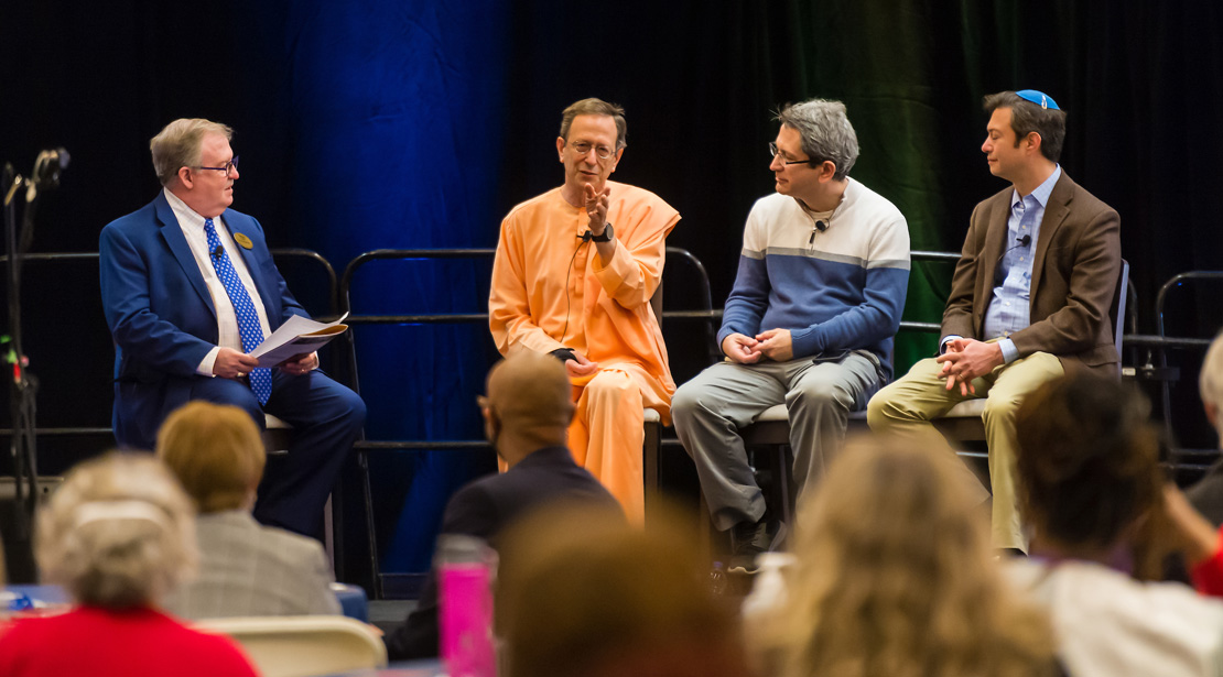Four light-skinned men sit on a stage, wearing: a blue suit and tie, a long orange robe with palm outstretched, a white and blue color-blocked sweater with gray slacks, and a brown jacket and tan pants with a blue yarmulke. Paul Stroble, Swami Nishpapananda, Elvir Mandzukic, and Rabbi Noah Arnow.