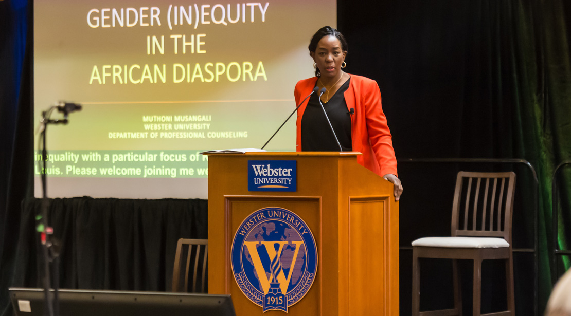 Muthoni Musangali, a dark-skinned woman, stands at a podium. Behind her, a large screen reads Gender (In)Equity in the African Diaspora.