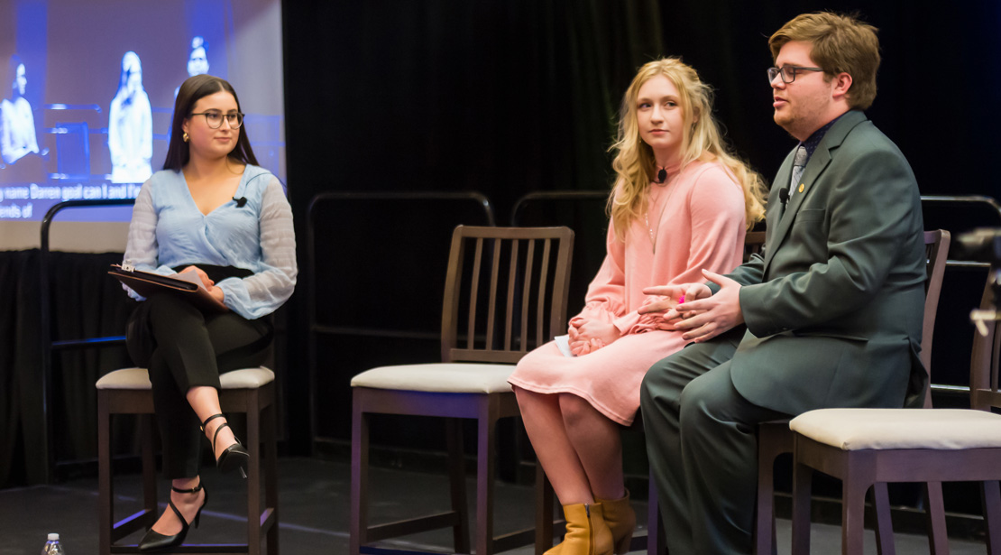 Three young light-skinned people sit on a stage: a woman wearing a blue long-sleeved shirt with black pants, a woman in a light pink knee-length dress, and a man wearing black-framed glasses and an all-gray suit. Katie Fields, Katarina Ausley, and Garret Dohlke.