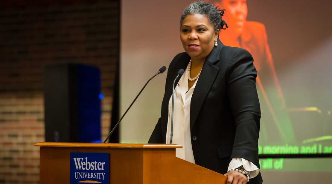 Rev. Traci Blackmon, an older medium-dark-skinned woman wearing a black jacket over a white shirt, stands behind a podium, hands gripping its sides.