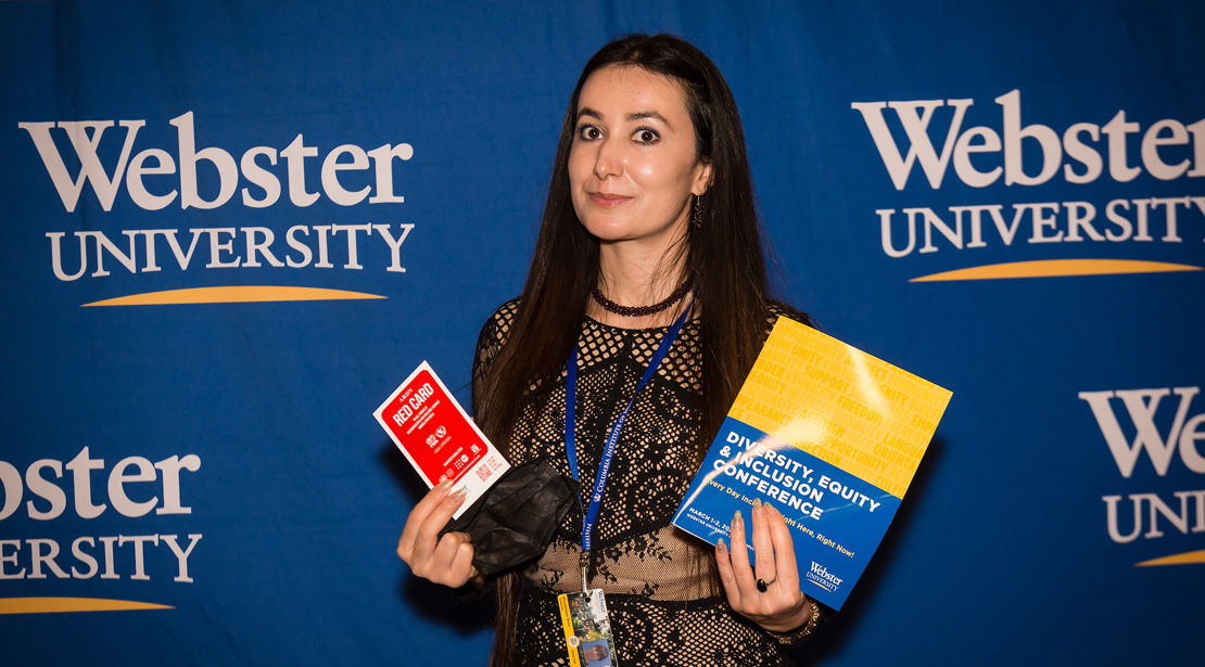 A medium-light-skinned woman with long, straight brown hair wearing a black and tan lace top and a lanyard around her neck stands in front of a blue backdrop with Webster University logo on it and holds up a Red Card and the Diversity, Equity & Inclusion Conference booklet.