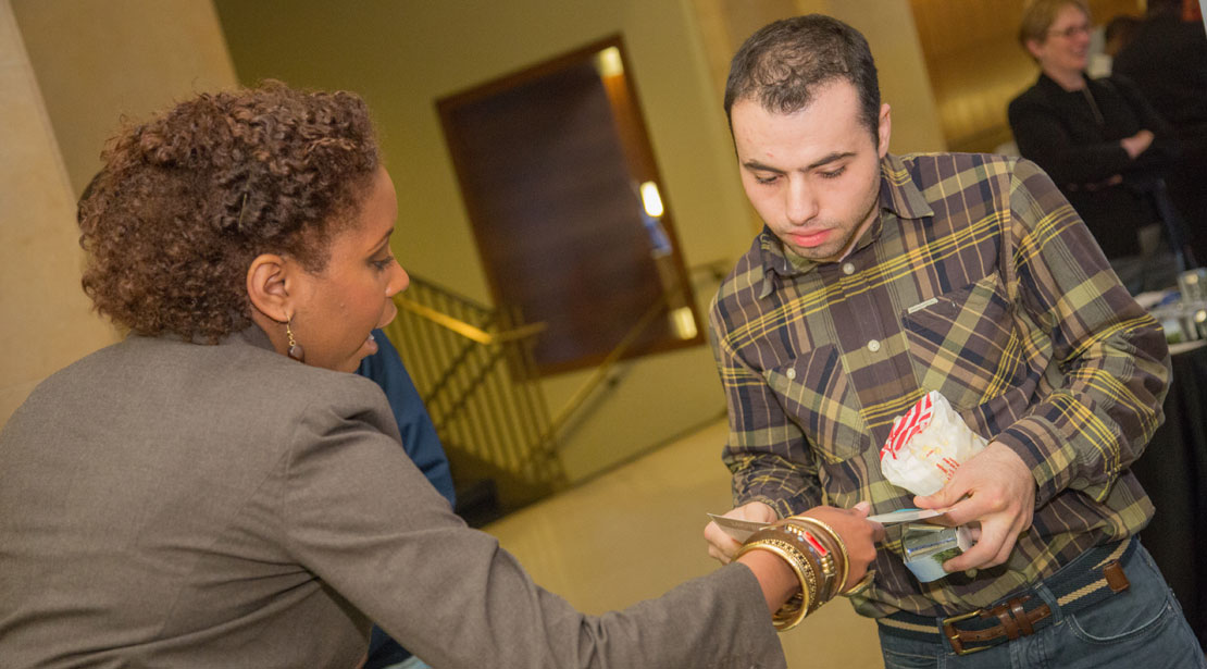 A medium-dark-skinned woman wearing a business suit hands a piece of paper to a light-skinned man wearing a plaid shirt.