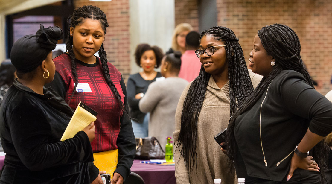 Four medium-dark-skinned women stand in a semi-circle, talking. One wears black with a black headwrap; the next wears a red-patterned shirt with black sleeves, her hair braided into two long plaits; the third wears a tan sweater, glasses, and long braids; the fourth wears all black with long braids tied back.