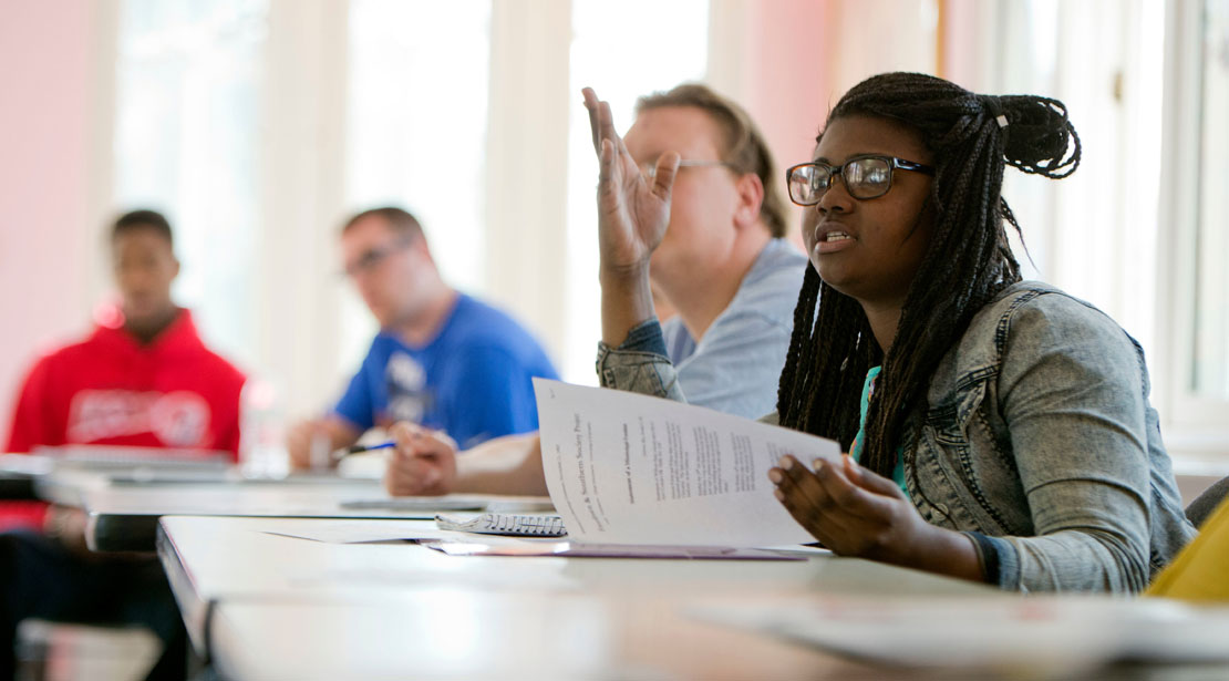 A young dark-skinned woman wearing glasses and a jeans jacket sits at a table with three other students. She is talking and holding a paper open; her other hand is raised.