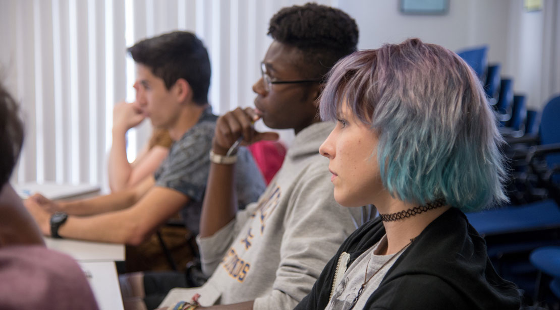 Three students - one light-skinned female with multi-colored hair, one dark-skinned male with hand to chin, one light-skinned male leaning forward - sit at a table and look towards the front of the room.