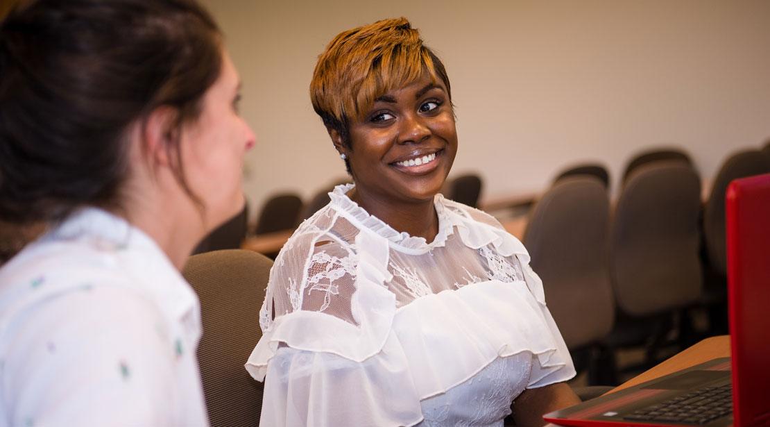 A smiling woman with medium-dark skin and short light-brown hair wearing a whole blouse sits at a desk in front of a laptop and looks at another woman with light skin tone wearing a white shirt.