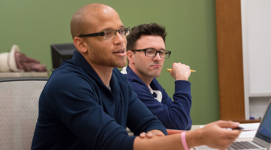 A bald, medium-dark-skinned man wearing glasses and a dark blue long-sleeved shirt talks while sitting next to a light-skinned man wearing glasses and a long-sleeved blue and white shirt who is holding a pencil.