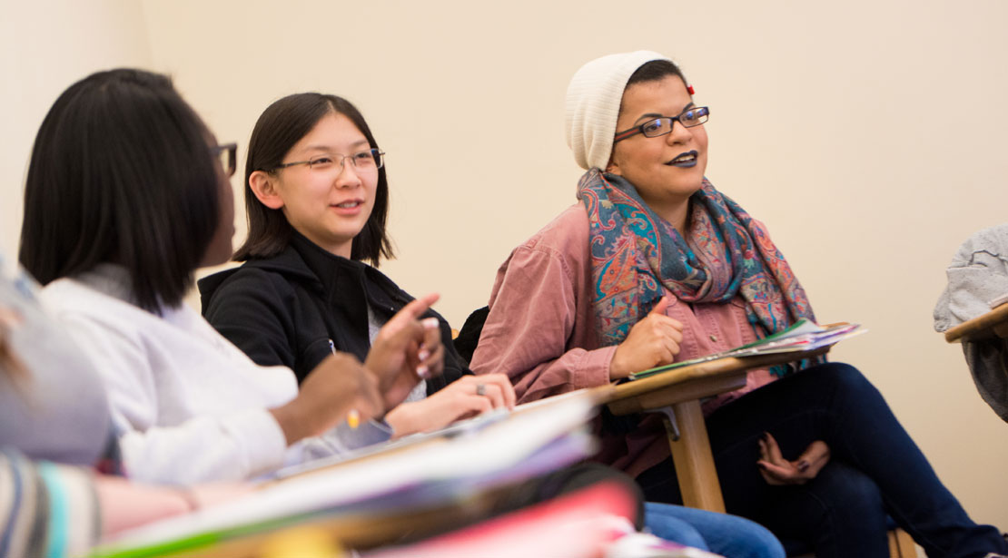 A diverse group of three women sits at desks smiling, talking, and gesturing.