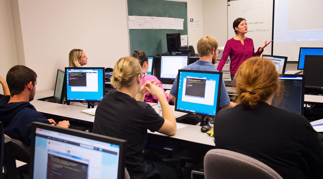 Professor stands talking and gesturing in front of a classroom with three long rows of tables with computers on each, students sitting at each computer showing computer code in small black content box on screen.