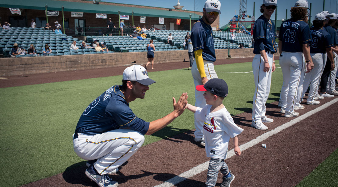 Webster baseball player at the end of a row of players standing on the baseball field squats to high five a young child.