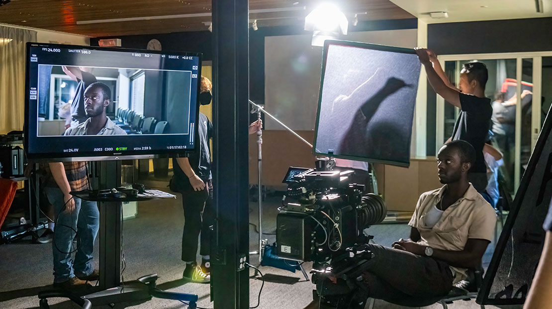 actor sits in chair and looks sideways at large video monitor as someone adjusts light filter behind him