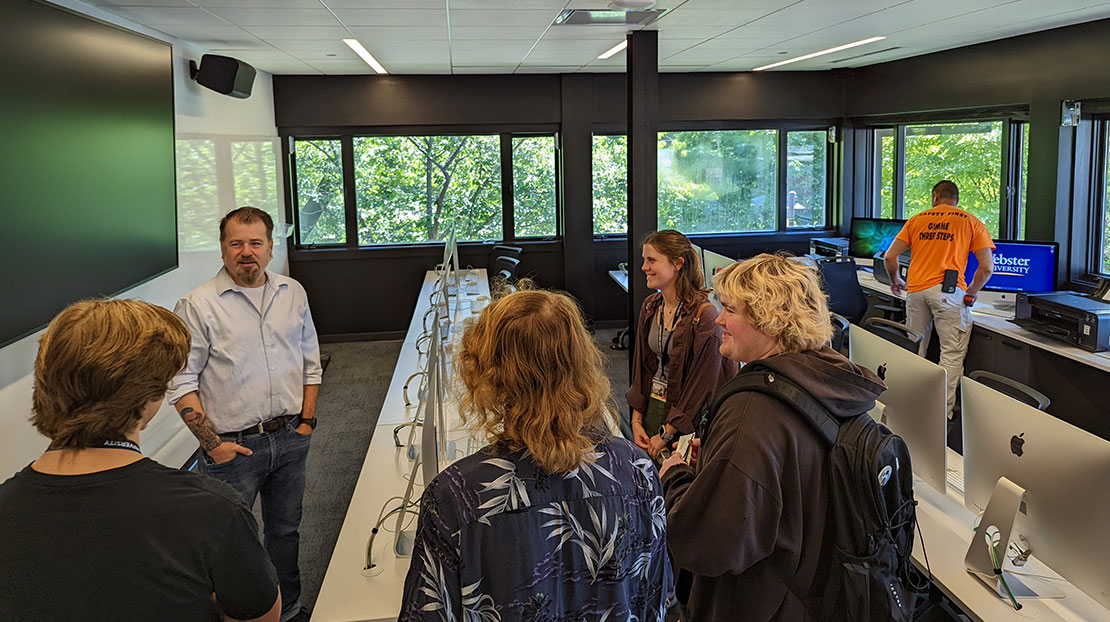 Professor AuBuchon talks with students in computer lab with rows of computers, large windows, and large TV monitor on wall
