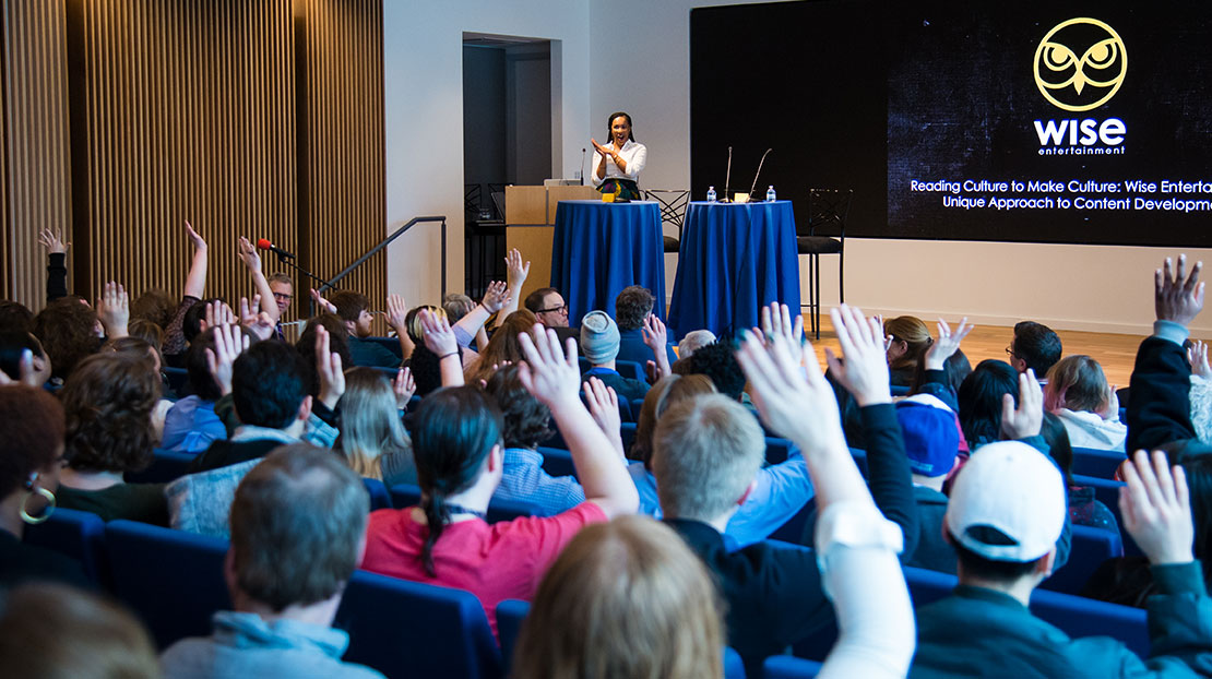 Auditorium of students with hands raised while presenter at podium claps