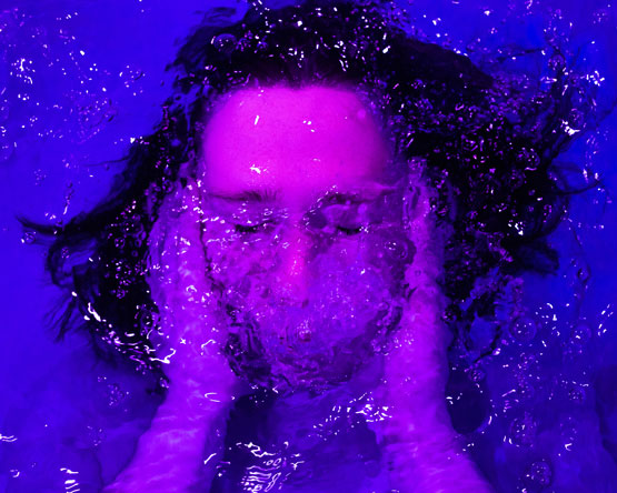 Underwater dark-haired person with eyes closed and hands on face, purple-blue water, bright lavender skin