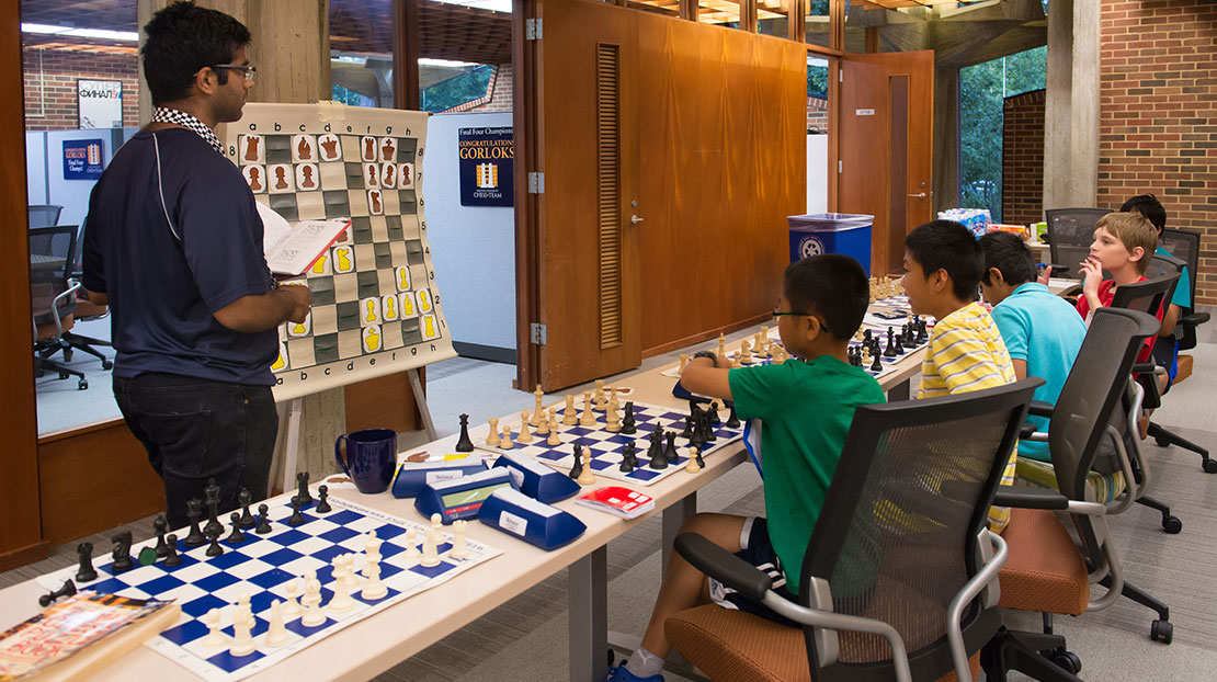 Group of young chess players at table with coach showing strategies on display chess board