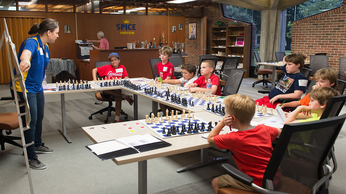 Group of young chess players at set of tables shaped in a U with coach showing strategies on display chess board