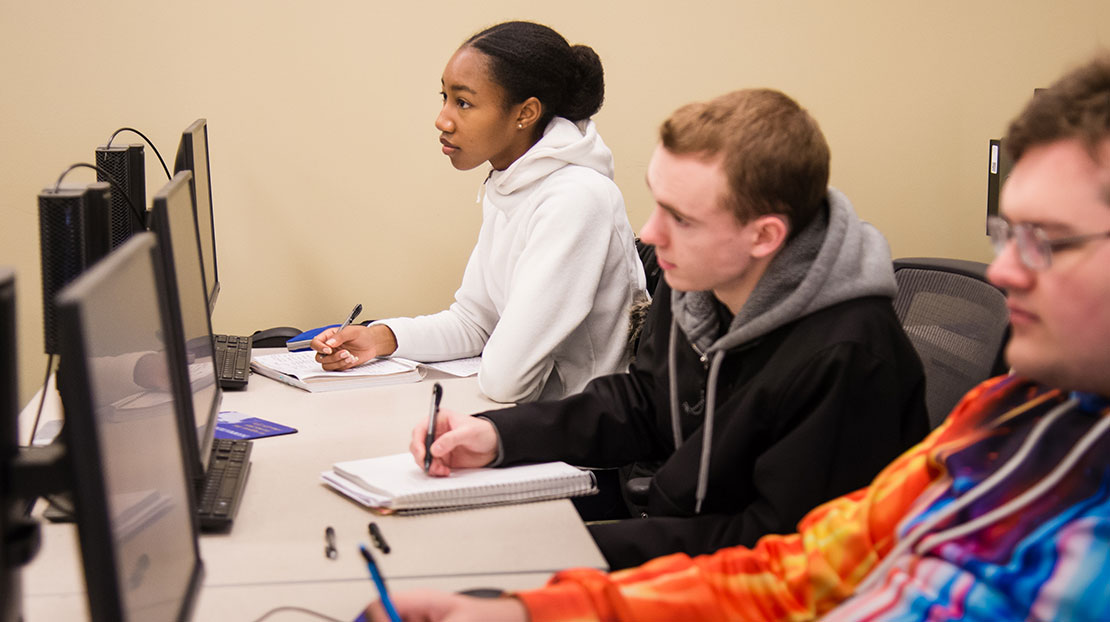 three students sit in front of computer monitors and take notes