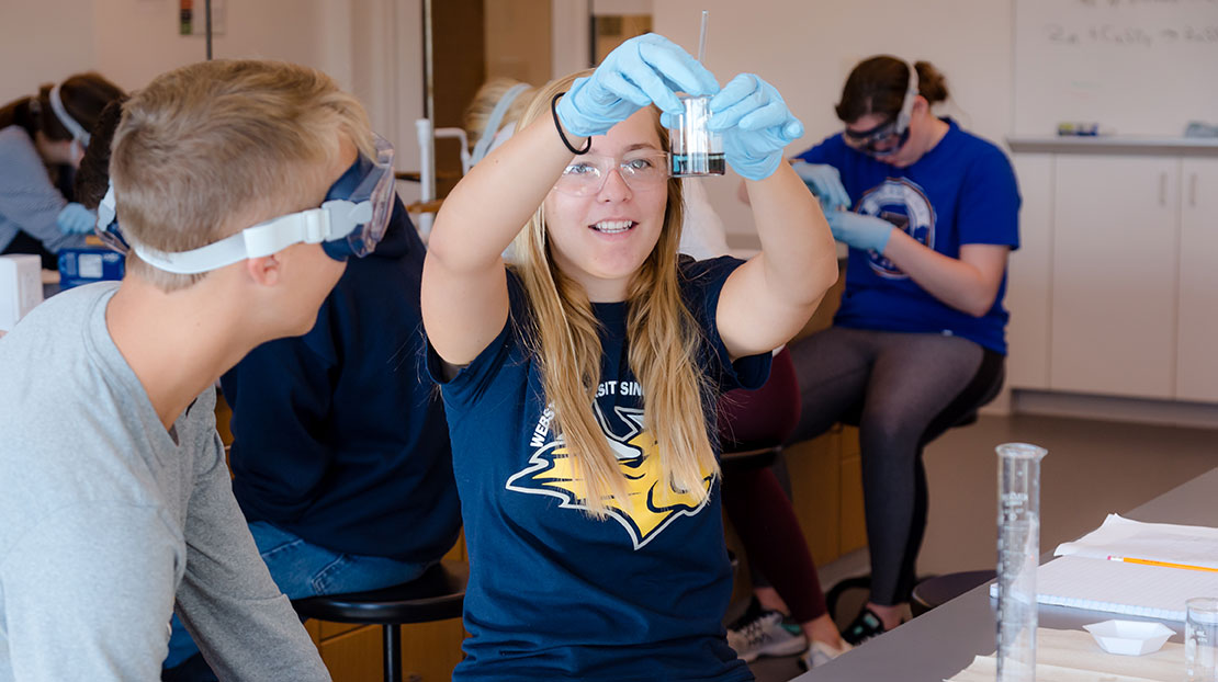 Two students look at chemical effect taking place in small beaker held by gloved student