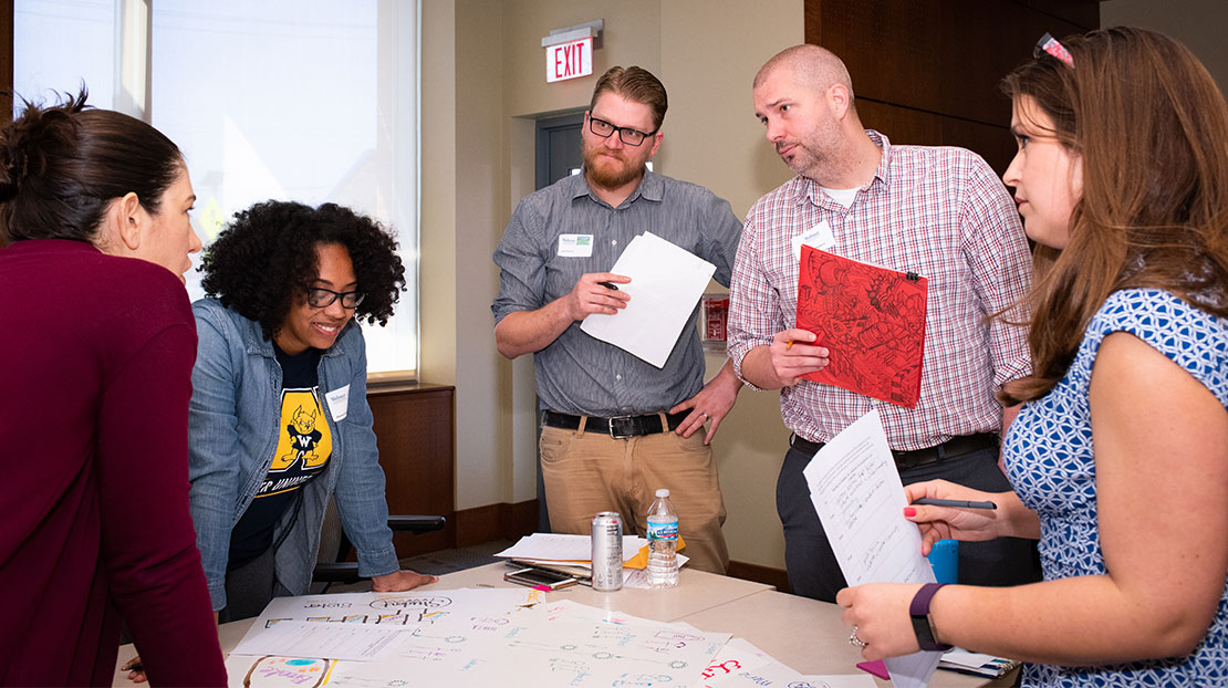 group of students work on poster board presentation while standing around a table