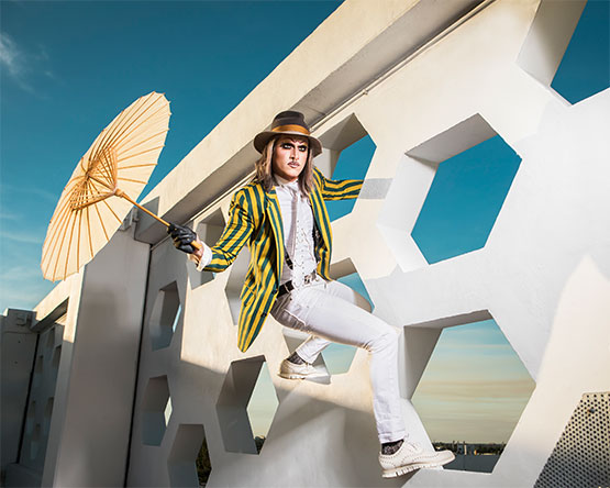 Person in white with yellow and blue striped jacket and brown fedora hat holding sun umbrella clings to wall with open hexagons