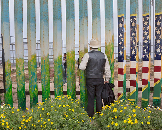 Man wearing grey suit vest and slacks and white cowboy hat looks through wall slats painted with American flag and field with yellow flowers.