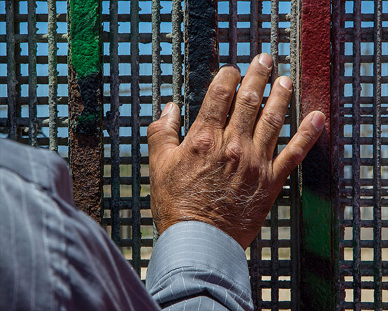 Close-up of a wrinkled man's hand against the painted border fence with cross-hatched fence behind it.