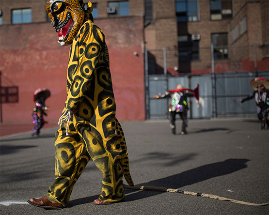 Person wearing stylized leopard mask and painted jumpsuit walks on asphalt in front of building.