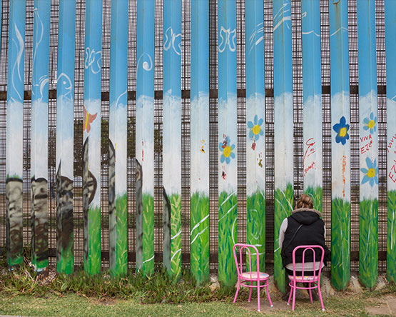 Woman in black vest sits on one of two pink chairs and looks through wall slats painted with field and flowers.