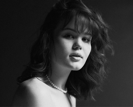 black and white of a woman with tousled hair and a pearl necklace, looking at the camera with a subtle expression