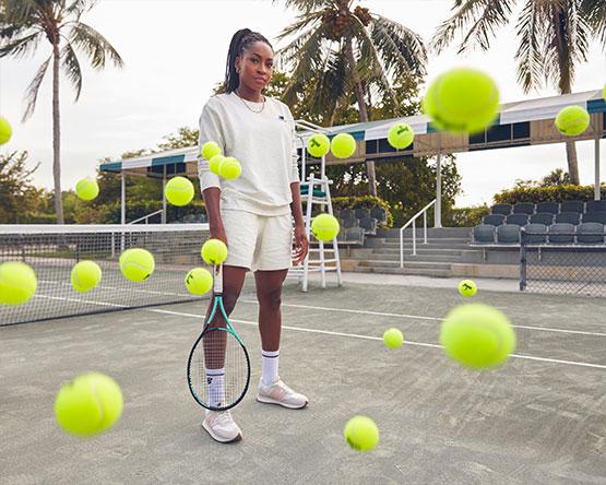 Coco Gauff stands holding tennis racket on court among bouncing tennis balls.