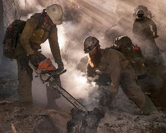 Firefighters work in haze and smoke to cut up smoldering wood.