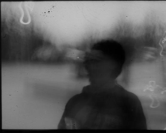 A grainy, black-and-white abstract photograph showing the blurry silhouette of a person against a pale, out-of-focus background.