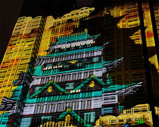 A vibrant light projection of a traditional Japanese castle layered over the facade of a modern skyscraper at night.