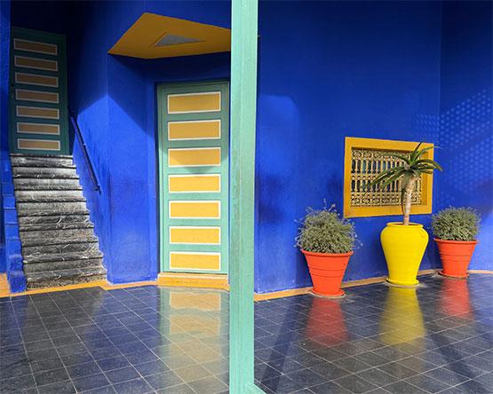 A bright courtyard with vivid blue walls, yellow-trimmed doors, and several colorful potted plants including a large yellow urn.