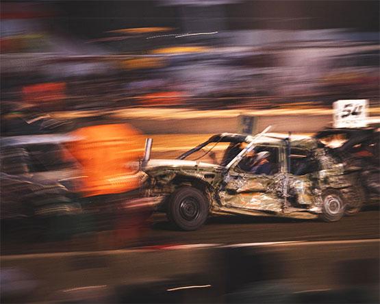 A motion-blurred action shot of a battered car competing in a demolition derby on a dirt track.