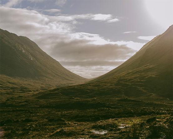 A wide, grassy mountain valley between two steep, shadowed slopes under a hazy, bright sky.