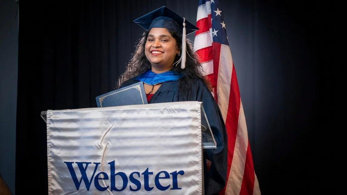 A person smiling while holding a diploma holder at the podium. 