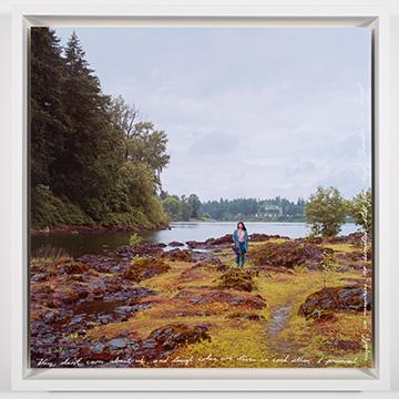 A full-frame view of a person wearing a blue coat standing on rocky, moss-covered ground near the shore of a calm lake surrounded by dense evergreen forests under a cloudy sky. Handwritten text runs along the bottom and right edge.