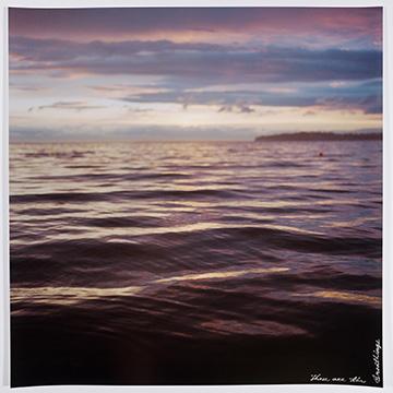 A close-up view of gentle water ripples reflecting the purple and gold light of a twilight sky, with a distant landmass on the horizon. Cursive text is written in the bottom-right corner.