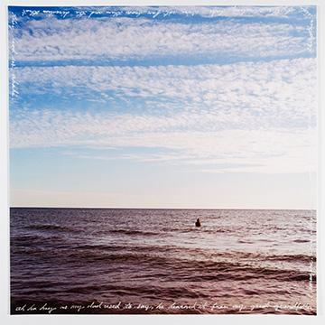 A wide view of a vast, rippling body of water under a big blue sky with soft, wispy clouds, with a lone swimmer submerged up to their shoulders near the horizon. Handwritten cursive text frames the edges of the print.