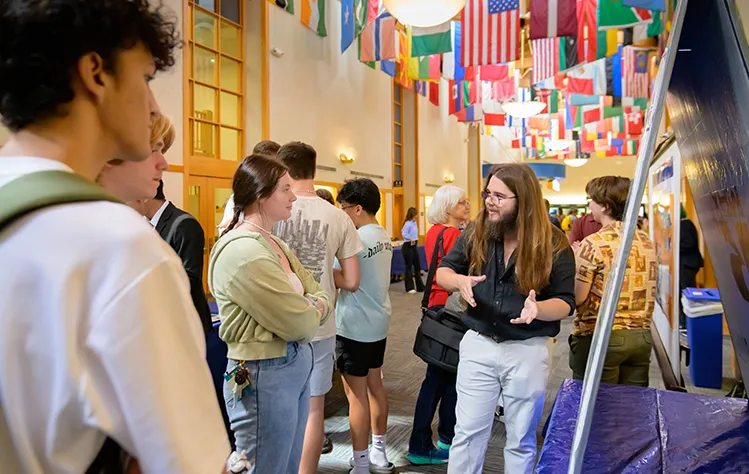 Student explains their project to a small group of peers in a hallway decorated with international flags.