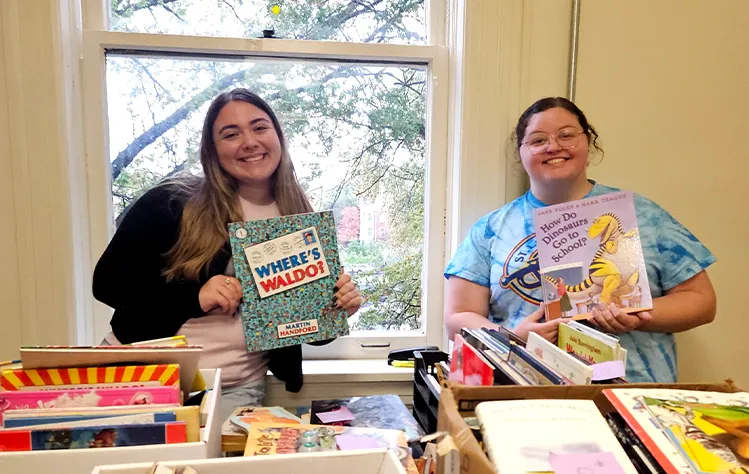 Two students smile while holding children’s books in front of a large window.