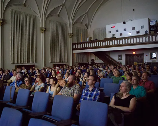 An audience seated in an auditorium with blue chairs, looking up toward the screen beneath a raised balcony and arched architectural details.