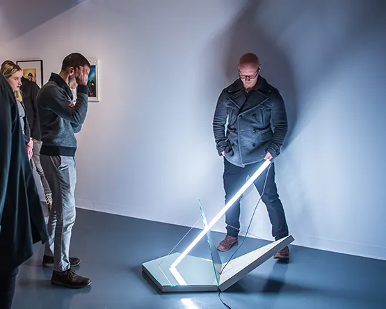 Visitors observing a lighted geometric floor sculpture composed of glass panels and a fluorescent tube light in the Hunt art gallery.