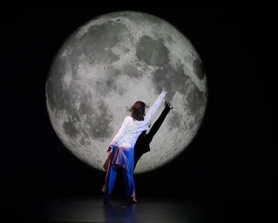 A Webster University dancer in a light-colored long shirt and loose pants seen from behind on a dark stage, reaching their left hand upward toward a large, brightly illuminated full moon projected in the background.