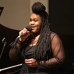 Webster University Jazz vocalist with braided hair styled in an updo holds a microphone and sings on stage in front of a black grand piano.