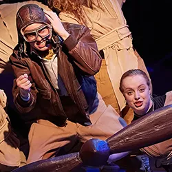 Webster Conservatory actor in bomber jacket flies plane composed of actors in tan outfits.