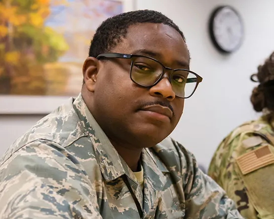 An active-duty service member wearing glasses and a camouflage uniform sitting in a classroom.